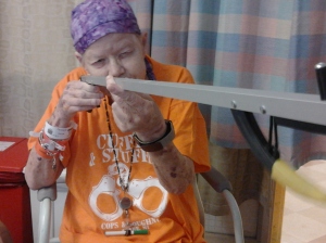 Mom in the hospital a few months before she died. She is wearing her favorite orange "Cuffed and Stuffed" t-shirt and using her grabber as if it is a rifle.